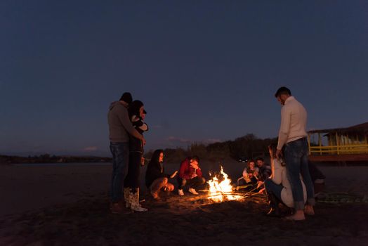 Happy Carefree Young Friends Having Fun And Drinking Beer By Bonefire On The Beach As The Sun Begins To Set