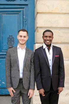 Two boys, caucasian and afro american, wearing suits standing near building. Concept of fashion and businessmen.