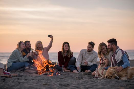 Happy Carefree Young Friends Having Fun And Drinking Beer By Bonefire On The Beach As The Sun Begins To Set