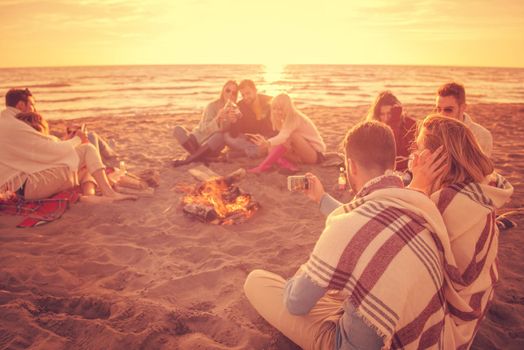 Happy Carefree Young Friends Having Fun And Drinking Beer By Bonefire On The Beach As The Sun Begins To Set filter