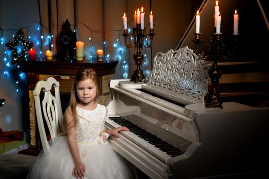 Beautiful little girl is playing on a white piano in a dark room by candlelight. The concept of Christmas and New Year, family happiness.