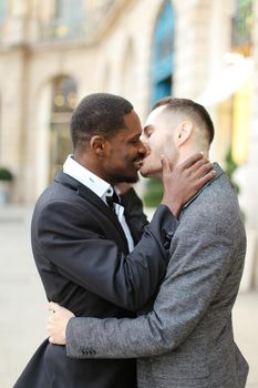 Afro american gay kissing caucasian boy outside, wearing suits. Concept of lgbt and same sex couple.