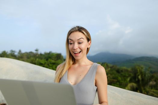 Young nice female person working with laptop in green mountains background, wearing grey shirt. Concept of modern technology, summet vacations and nature.