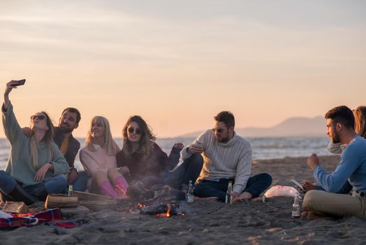 Happy Carefree Young Friends Having Fun And Drinking Beer By Bonefire On The Beach As The Sun Begins To Set