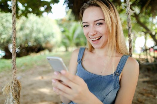 Young smiling female person riding swing and using smatrphone, sand and tree in background. Concept of summer vacations and modern technology.