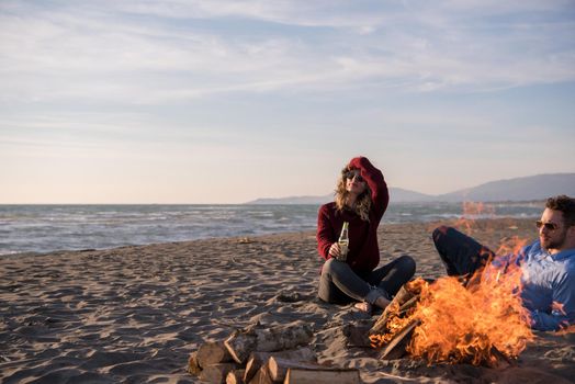 Young Couple Relaxing By The Fire, Drinking A Beer Or A Drink From The Bottle on the beach at autumn day