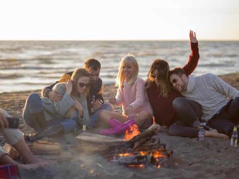 Happy Carefree Young Friends Having Fun And Drinking Beer By Bonefire On The Beach As The Sun Begins To Set