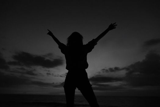 Black silhouette of female person with raised hands on grey sunset sky background in Bali. Concept of last minute tours to tropical resort, summer vacations and gladden tourist.