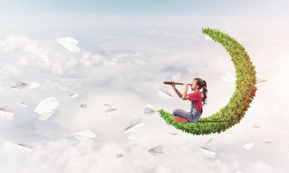 Cute kid girl on green moon in day sky looking in spyglass