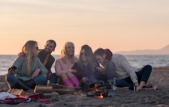 Happy Carefree Young Friends Having Fun And Drinking Beer By Bonefire On The Beach As The Sun Begins To Set