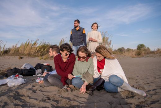 Happy Carefree Young Friends Having Fun And Drinking Beer By Bonefire On The Beach As The Sun Begins To Set