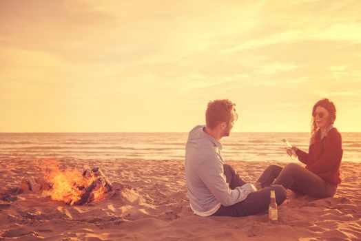 Young Couple Relaxing By The Fire, Drinking A Beer Or A Drink From The Bottle on the beach at autumn day filter