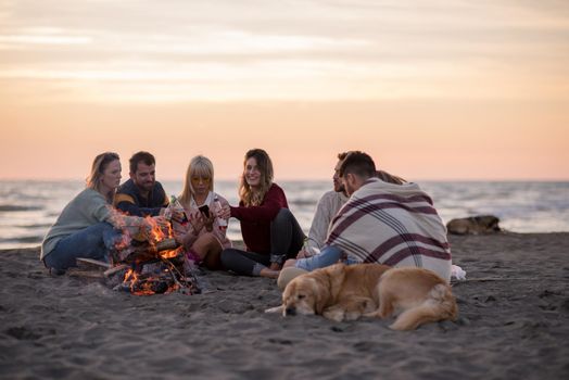 Happy Carefree Young Friends Having Fun And Drinking Beer By Bonefire On The Beach As The Sun Begins To Set