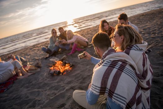 Happy Carefree Young Friends Having Fun And Drinking Beer By Bonefire On The Beach As The Sun Begins To Set