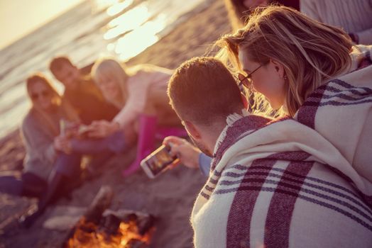 Happy Carefree Young Friends Having Fun And Drinking Beer By Bonefire On The Beach As The Sun Begins To Set filter