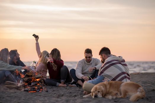 Happy Carefree Young Friends Having Fun And Drinking Beer By Bonefire On The Beach As The Sun Begins To Set