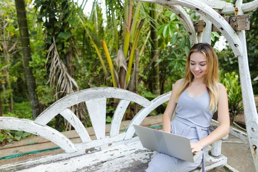 Young girl sitting on swing and using laptop, palms in background. Concept of exotic summer vacations and modern technology.