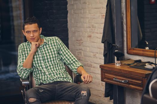 Handsome young man in shirt and jeans looking at the camera and sitting in a chair at the barbershop. He looks confident