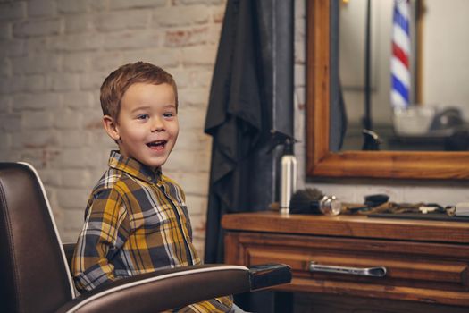 Portrait of a stylish little boy dressed in shirt and jeans sitting in a chair against the barber's workplace, he smiles and posing before camera