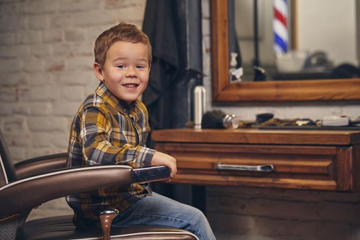 Portrait of a stylish little boy dressed in shirt and jeans sitting in a chair against the barber's workplace, he smiles and posing before camera