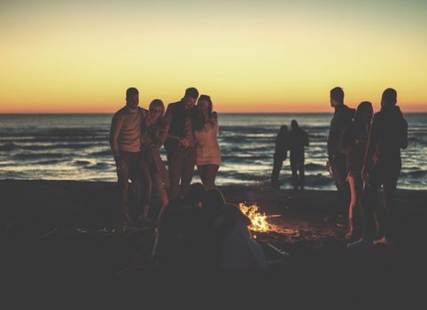 Happy Carefree Young Friends Having Fun And Drinking Beer By Bonefire On The Beach As The Sun Begins To Set colored filter