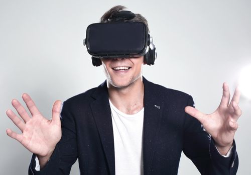 Young handsome businessman in black suit wearing virtual reality goggles, isolated on gray background