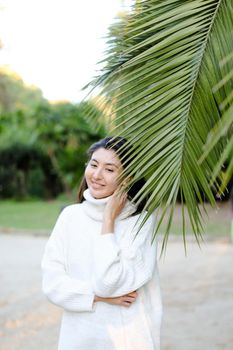 Korean nice woman in white sweater standing near palm leaf. Concept of asian beauty and female person on tropical resort.