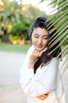 Portrait of chinese woman wearing white sweater standing near palm leaf. Concept of asian beauty and female person.