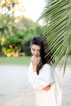 Korean dreaming woman in white sweater standing near palm leaf. Concept of asian beauty and female person on tropical resort.