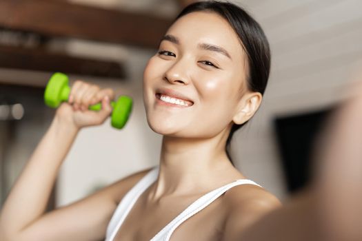 Portrait of smiling asian fitness instructor taking selfie with dumbbell. Athletic woman making photo on mobile phone with sport gear, workout at home.