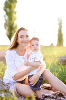 Young nice woman sitting with little baby on plaid, grass on background. Concept of picnic, motherhood and nature.