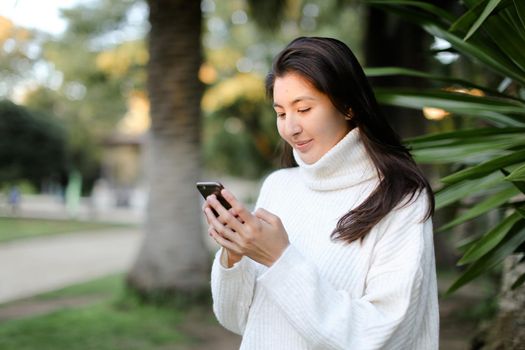 Korean girl using smartphone and walking in tropical park. Concept of modern technology and nature.