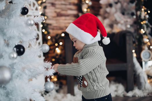 Pretty caucasian boy with a red Christmas hat decorates a big New Year's white christmas tree