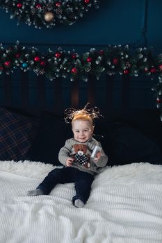 Portrait of lovely little toddler sitting on bed with illuminated garland on his head and smiling.