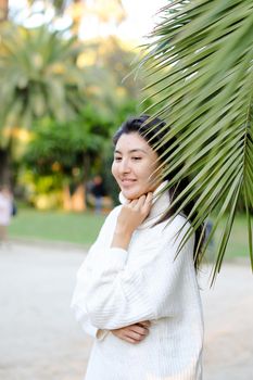 Chinese beautiful woman in white sweater standing near palm leaf. Concept of asian beauty and female person on tropical resort.