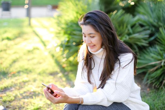 Chinese female student sitting in park and typing by smartphone. Concept of modern technology and social networks.