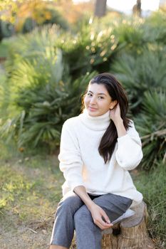 Chinese pretty woman sitting on stump in park and wearing white sweater. Concept of asian female person.