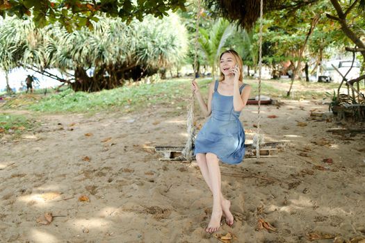 Young european woman riding swing and talking by smatrphone, sand and tree in background. Concept of summer vacations on tropical and exotic resort, modern technology.