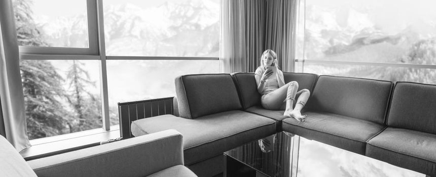 Young beautiful girl enjoying music through headphones, laying on sofa near the window at home