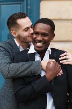 Two boys, caucasian and afro american, wearing suits standing near building and hugging. Concept of gays and lgbt.