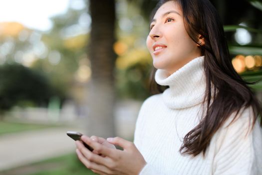 Chinese cute girl using smartphone and walking in tropical park. Concept of modern technology and nature.