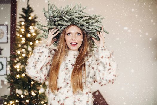 Waist-up photo of excited red-haired lady wearing a crown of fir branches in a swarm of snowflakes