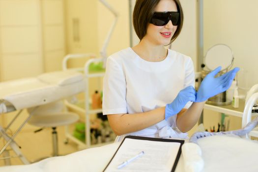 Young cosmetologist wearing special purpose glasses and latex gloves near permanent makeup device. Concept of beauty cosmetology salon and expensive high quality equipment.