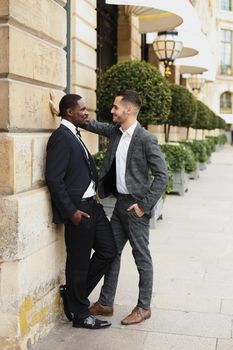 Afro american and european gays standing near building and wearing suits and talking outside. Concept of lgbt and walking in city.