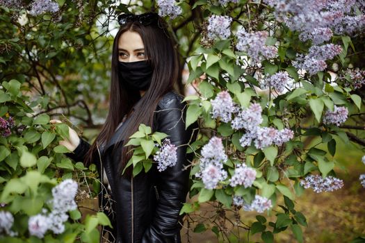 Girl in a medical mask on the background of a blossoming lilac in the park. Black mask. Protection against virus, flu. Coronavirus protection. epidemic of coronavirus. Spring allergy.