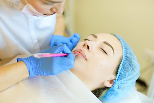 Skilled cosmetologist shaping lips with pencil for young woman before microblading. Concept of permanent makeup artist and beauty salon.