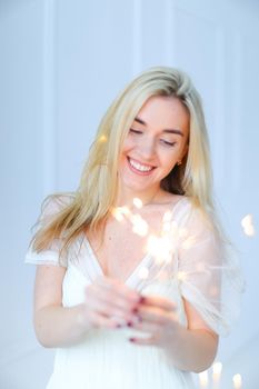 Blonde beautiful girl keeping bengal light and wearing white dress. Concept of happiness and celebrating holidays.