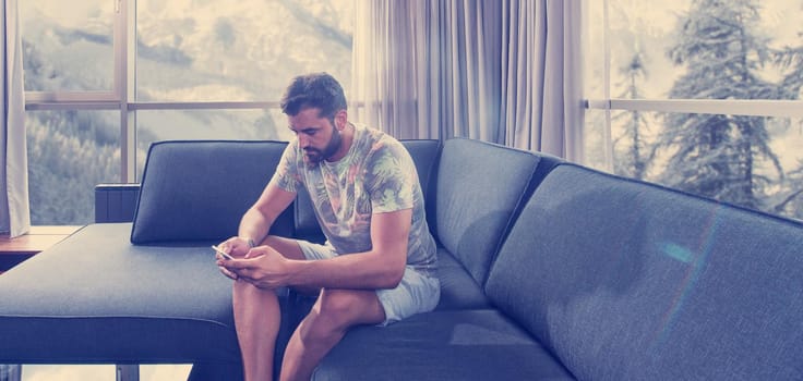 young man sitting on sofa and using a mobile phone  near the window at home