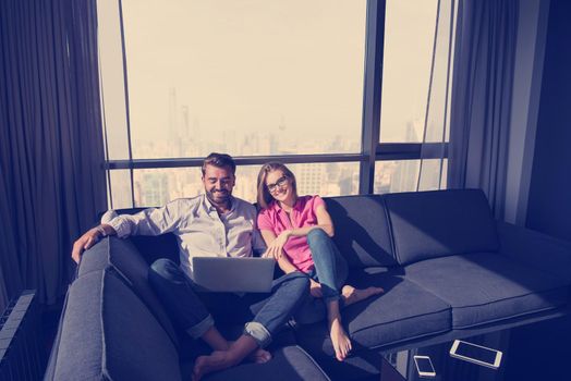 Young couple relaxing at  home using laptop computers reading in the living room near the window on the sofa couch.