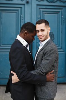 Afro american handsome man hugging caucasian guy in door background, wearing suit. Concept of same sex couple and gays.